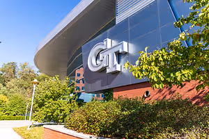 The Georgia Tech campus features a modern building with a curved roof and a large sign that reads GT, surrounded by greenery and trees under a clear blue sky.