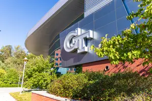 The Georgia Tech campus features a modern building with a curved roof and a large sign that reads GT, surrounded by greenery and trees under a clear blue sky.