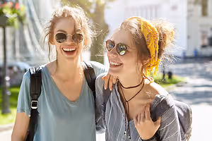 Two women are smiling and posing for a photograph on the road near the street light and streetlights. They are wearing sunglasses and carrying backpacks. The woman on the left is carrying a handbag, and the other is wearing a headband and a necklace. Behind them is a building with glass windows and a lamp on the side. The area is surrounded by trees and grass.