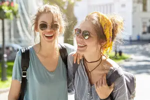 Two women are smiling and posing for a photograph on the road near the street light and streetlights. They are wearing sunglasses and carrying backpacks. The woman on the left is carrying a handbag, and the other is wearing a headband and a necklace. Behind them is a building with glass windows and a lamp on the side. The area is surrounded by trees and grass.