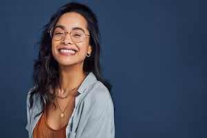 A woman with black hair wearing glasses and necklaces is smiling and posing for a photo against a blue background.