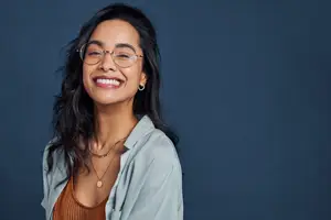 A woman with black hair wearing glasses and necklaces is smiling and posing for a photo against a blue background.