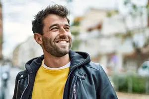 A man with a beard is smiling while standing on a street, wearing a yellow shirt and a black jacket with a zipper. Behind him, there are some blurry buildings, cars, and plants, and the sky is covered with clouds.