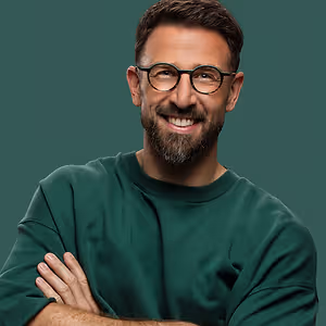 A smiling man wearing glasses and a green shirt is standing against a green background, possibly in a studio.