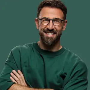 A smiling man wearing glasses and a green shirt is standing against a green background, possibly in a studio.
