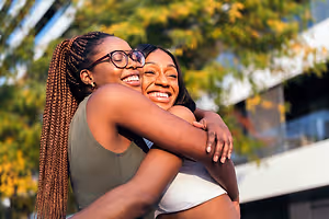 Two women are hugging each other outside. They are smiling and seem to be happy. They are standing in front of a building with a balcony and trees in the background.