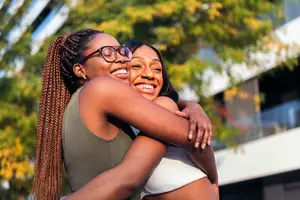 Two women are hugging each other outside. They are smiling and seem to be happy. They are standing in front of a building with a balcony and trees in the background.