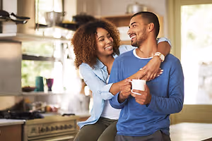 A smiling couple sharing a moment in a kitchen, with one person holding a cup and the other person embracing them