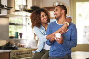A smiling couple sharing a moment in a kitchen, with one person holding a cup and the other person embracing them