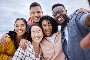 Five people are standing close together, smiling and posing for a photo on a beach, with one of them wearing glasses and a necklace. Behind them, there are blurry views of the beach, the ocean, and the sky.