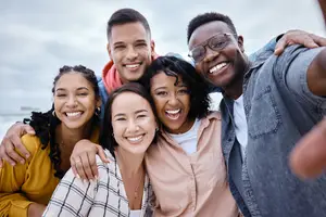 Five people are standing close together, smiling and posing for a photo on a beach, with one of them wearing glasses and a necklace. Behind them, there are blurry views of the beach, the ocean, and the sky.
