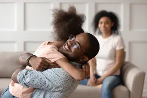 A man with glasses holds a child on his back while a woman sits on the couch behind them