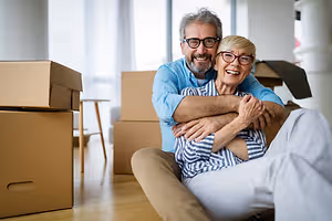Senior couple moving into their new home, sitting on the floor and smiling with boxes around them.