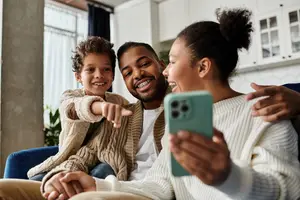 A family sitting on a couch, the man with braces is smiling, and the woman is laughing while pointing at the camera, and the boy is smiling and pointing at something on the phone, and the boy is wearing a sweater, and the man is wearing a white shirt, and the woman is wearing a white sweater, and they are sitting in a living room with white cabinets, a window with curtains, and a plant