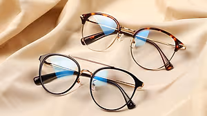 A pair of glasses with blue lenses and black frames is placed on a cream fabric cloth. The lenses reflect light and have a blue hue. The glasses are probably displayed in a store or a shop.