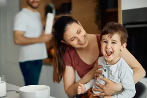 A woman holds a laughing child at a table with a glass of milk and a man holding a cup behind them.