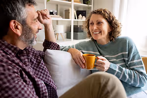 A man and woman sitting on a couch, the man resting his head on his hand and the woman holding a cup and smiling