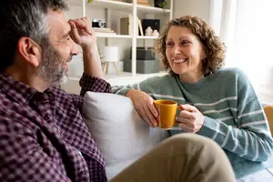 A man and woman sitting on a couch, the man resting his head on his hand and the woman holding a cup and smiling
