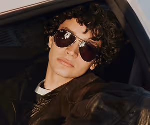 A young man with curly hair is sitting inside a car and wearing sunglasses and a leather jacket.