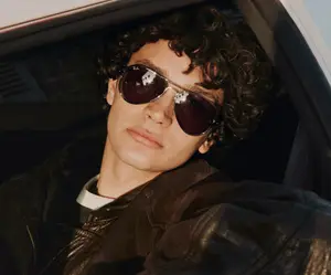 A young man with curly hair is sitting inside a car and wearing sunglasses and a leather jacket.