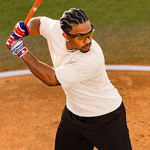 A man wearing a white t-shirt, black pants, and sunglasses is hitting a baseball with a baseball bat on a baseball field