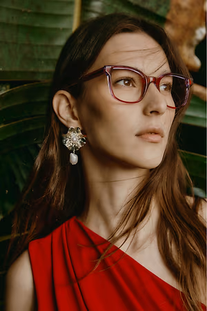 A woman with long brown hair and red glasses is wearing a red dress and earrings.