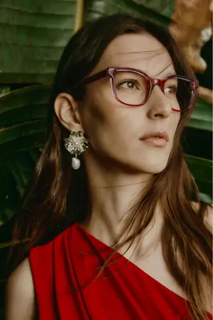A woman with long brown hair and red glasses is wearing a red dress and earrings.