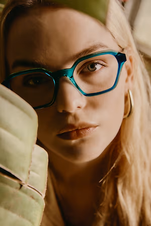 A close-up portrait of a woman with long blonde hair, wearing blue-rimmed glasses and holding a green leaf.