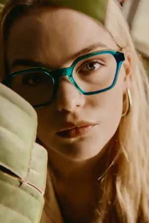 A close-up portrait of a woman with long blonde hair, wearing blue-rimmed glasses and holding a green leaf.