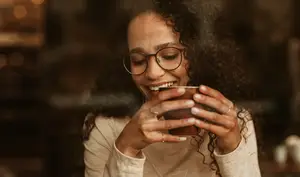 A smiling woman wearing glasses and holding a cup of coffee, looking at the camera with a warm smile.