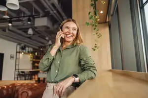A woman in a green shirt stands by a window and talks on a cell phone.