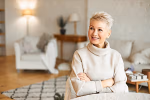 An adult woman with short hair smiles and poses for a photo in a room with a carpet, a desk, and a wooden table with a lamp and a plant