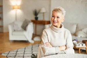 An adult woman with short hair smiles and poses for a photo in a room with a carpet, a desk, and a wooden table with a lamp and a plant