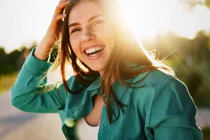 A woman in a green shirt smiles and laughs outside in the sunlight