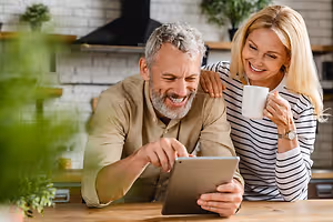An elderly couple sits in the kitchen, the man pointing at a tablet, while the woman holds a coffee mug and smiles at him