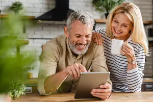 An elderly couple sits in the kitchen, the man pointing at a tablet, while the woman holds a coffee mug and smiles at him