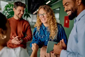 A group of four people in an office or meeting room, smiling and shaking hands, one holding a tablet, with a green plant and red fire extinguisher in the background.