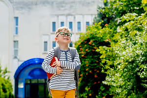 A boy with glasses carrying a book and a backpack is standing outside a building, smiling, and looking up at the sky.
