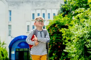 A boy with glasses carrying a book and a backpack is standing outside a building, smiling, and looking up at the sky.