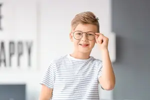 A boy in a white shirt and glasses is smiling and adjusting his glasses in a room with a white wall and the word Happy on it.