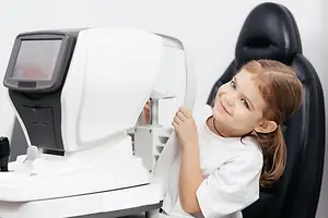 A young girl is smiling and holding a white machine with a screen, probably an ophthalmoscope, and is possibly in an eye clinic.