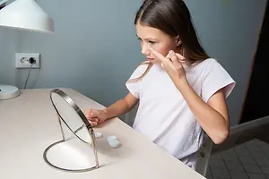 A girl is sitting in front of a desk, touching her face with one hand while looking at her reflection in a mirror. She is wearing a white shirt and has long hair. On the desk, there is a lamp, a power outlet, and a mirror stand with a mirror on it.