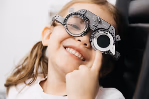 A young girl wearing a pair of eyeglasses is smiling and pointing her finger to the camera.