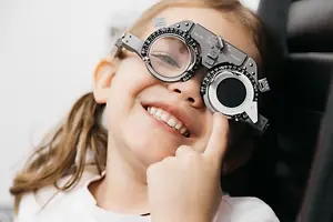A young girl wearing a pair of eyeglasses is smiling and pointing her finger to the camera.