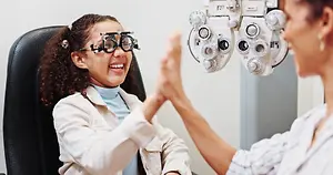 A young girl with glasses on is smiling at the camera while a woman looks at her with an eye exam machine behind them