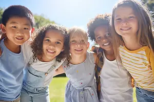 Five smiling children standing together on a grassy field with trees in the background