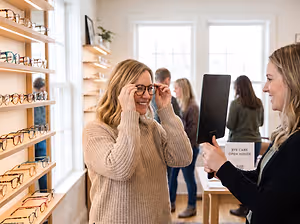 A woman holding a black object and showing it to another woman in a shop with shelves full of glasses.