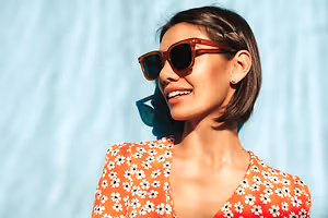 A woman with short hair is smiling and posing for a photo while wearing sunglasses and an orange floral blouse in front of a blue wall.