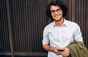 portrait of smiling young man holding phone and jacket in front of wooden fence