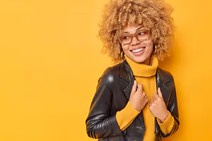 Portrait of a smiling woman with curly hair, glasses, and a yellow turtleneck, standing against a yellow background.